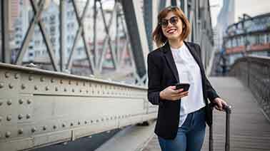 Eine Frau im Businesslook steht auf einer Brücke, hat ihr Handy in der Hand und lächelt in die Kamera