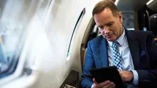 Ein Mann im Anzug sitzt in der Business Class eines Flugzeuges und tippt auf einem Tablet